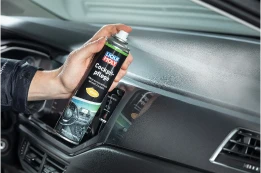 Hand hält Cockpit-Pflegespray und reinigt Armaturenbrett im Auto, Kunststoffoberfläche im Vordergrund.